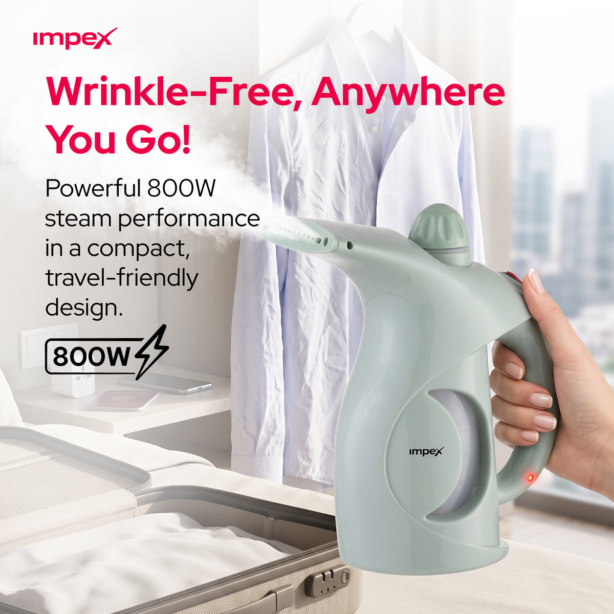 Impex handheld steam iron shown being held by a hand in front of a blurred bedroom scene with a white shirt and suitcase. The product is light sage green with a gray handle and a small red power button.